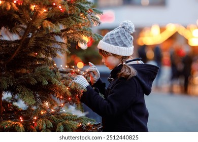 Little cute kid girl having fun on traditional Christmas market during strong snowfall. Happy child enjoying traditional family market in Germany. Schoolgirl standing by illuminated xmas tree. - Powered by Shutterstock