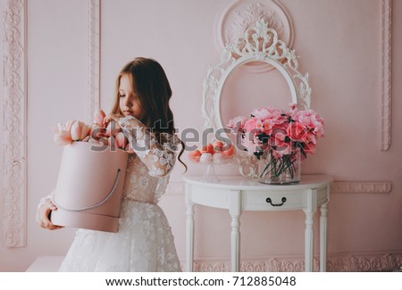 Little cute princess girl in white dress is holding flowers in a pink room.