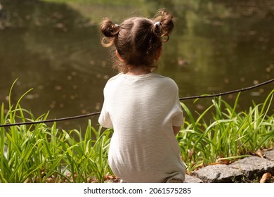Little cute redhead girl with ponytails sits on the shore of a pond with her back to the camera