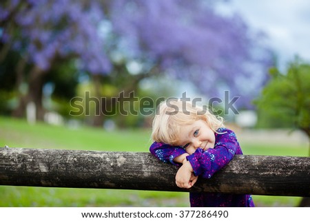 Little cute smiling girl in purple shirt is standing next to fence against blooming purple trees background