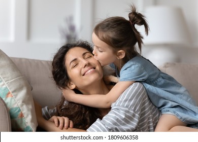 La pequeña hija levanta gentilmente abrazos a la madre en la mejilla mostrando amor y amor expreso descansando en el sofá en casa. Feliz familia, placer, mamá, felicitaciones por el día de la madre, dulce momento juntos concepto Foto de stock