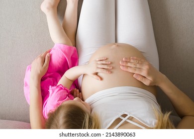 Little Daughter Hand Touching Pregnant Mother Naked Big Belly And Feeling Baby Move. Cute Moment. Waiting Future Brother Or Sister. Sitting Together On Sofa At Home. Emotional Loving Pregnancy Time.