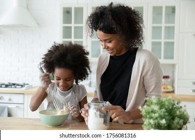 Little daughter helper and African mother cooking together in kitchen, kid girl preparing cake or pancakes enjoy process mixes dough using whisk, teach and upbringing, healthy home food, hobby concept - Shutterstock ID 1756081316