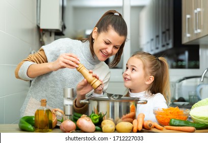 Little daughter helping cooking soup and mother add pepper to pan - Powered by Shutterstock