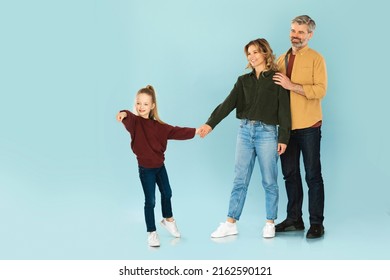 Little Daughter Holding Hands With Parents And Pointing Finger Aside Advertising Family Offer Standing Posing On Blue Studio Background. Look There Concept. Full Length Shot