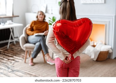 Little daughter holds red heart shaped balloon behind her back, a gift for mom at home. Mothers Day celebration.