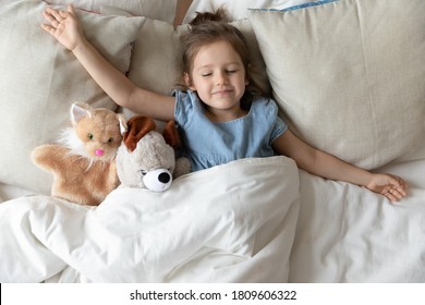 Little Daughter With Outstretched Hands Lying In Bed With Fluffy Stuffed Toys Animal Friends Cat And Teddy Bear Sleeping In Cozy Room. Kid Girl Covered With Blanket Enjoy Healthy Night Sleep Top View