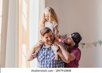 Little daughter sitting on her daddy's neck. Portrait of happy gay couple and their beautiful child indoors.