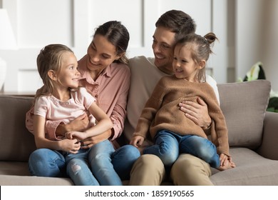 Little daughters sit on parents laps talking together resting on couch in living room. Married couple spend time communicating with 2 cute kid girls on weekend at home. Family bonds, adoption concept