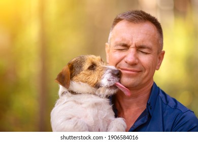 Little Dog Licks Human Face. Man With His Dog Playing In The Park. Owner Hugs His Pet.