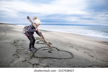 A little dreamy dark-haired girl in warm clothes and a white hat with a large hiking backpack walks on my empty sea sandy beach, and draws children's drawings and symbols on the wet sand