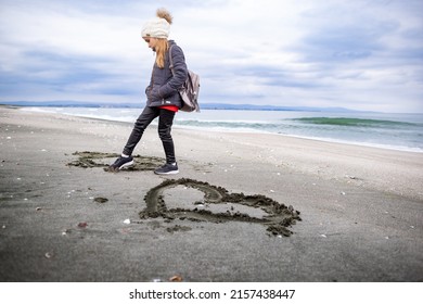 A little dreamy dark-haired girl in warm clothes and a white hat with a large hiking backpack walks on my empty sea sandy beach, and draws children's drawings and symbols on the wet sand