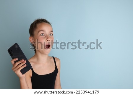 Little emotional teenage girl in a black t-shirt 11, 12 years old on an isolated blue background with a smartphone in her hands. Children's studio portrait. Place text to copy space for caption.