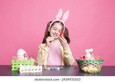Little enthusiastic child drawing colorful designs and painting eggs with watercolors to prepare for the Easter festivities, pink backdrop. Young small girl relaxing as she creates art at table. - Powered by Shutterstock