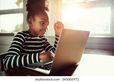 little ethnic girl sitting at home at a table working on her homework on a laptop computer reading the screen with a thoughtful expression - Powered by Shutterstock