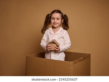 Little European girl, adorable kid in pajamas holds a craft cardboard house model being inside a box, against beige background with copy space for ads. The concept of investment, housing