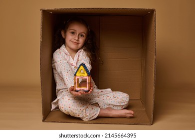 Little European girl holding a house built from magnetic square and triangle construction blocks, sitting inside a cardboard box and cutely smiling looking at camera, isolated over beige background