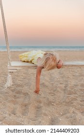Little Fair-haired Girl Lies On Wooden Swing. Toddler Pushes In Rope Swing On The Beach In Summer Evening. Bored Child.