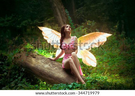little fairy in pink dress, with transparent golden pink glowing wings, fashion creative color, fantastic shooting Forest nymph butterfly sits on a log. Fairy goddess of nature harmony. 
