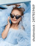 Little fashion kid girl 5-6 years old in blue poofy dress and sunglasses talk by mobile cell phone. Top view kid portrait