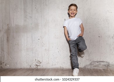 Little fashionable boy posing in front of gray concrete wall. Portrait of smiling child in white T-shirt and gray trousers. Concept of style and fashion for children.