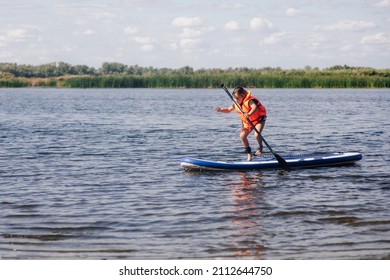 Little female child standing on paddle board on clue lake trying to keep balance on feet wearing orange life jacket. Active holidays full of adventures. Inculcation of love for sports from childhood. - Shutterstock ID 2112644750