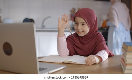 The Little Female Student Wearing A Headscarf Is Watching The Lessons On The Internet With A Computer Video Call And Working From Home. She Asks Questions To Her Teacher And Takes Notes. 