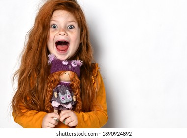 Little five-year-old red-haired girl happy screaming yelling. Child kid holds a red hair doll in her hands in an orange sweater on white background