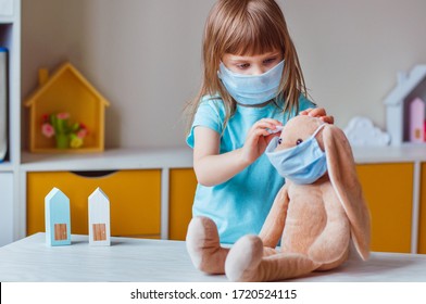 Little Four Years Old Girl In The Face Mask Wearing Her Rabbit Soft Toy In The Same Mask In The Kids Room. Social Distance Stay At Home During Covid-19 Pandemic Concept.