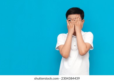 little frightened asian boy in white t-shirt covers his face with his hands and peeks on blue background, korean child is shy and hides on isolated background