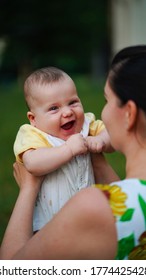 Little Funny Baby On Arms Of Mother Laughing With Wide Open Mouth Outdoors