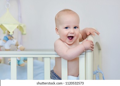 Little funny boy standing in baby bed and laughing. Smiling adorable child on feet in playpen. Kid portrait at nursery. Happy childhood concept