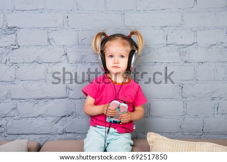 A little funny girl of two years is sitting on the couch, listening to music on headphones put on her head. Holds a smartphone in a bag in the form of a rabbit.