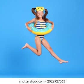 Little funny happy girl in swimsuit and goggles on her head jumping up in air with inflatable ring around waist, isolated over blue studio background. Summertime, vacation an school holidays concept