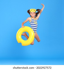 Little funny happy girl in swimsuit and goggles on her head jumping up in air with inflatable ring around waist, isolated over blue studio background. Summertime, vacation an school holidays concept