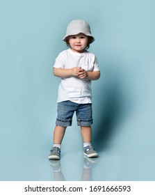 Little ginger toddler boy or girl in white t-shirt, gray hat, socks and shoes, denim shorts. Child is smiling while posing on blue background. Childhood, fashion, advertising. Full length, copy space