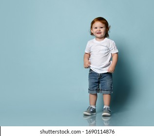 Little ginger toddler male or female in white t-shirt, socks and shoes, denim shorts. Child is smiling while posing on blue background. Childhood, fashion, advertising. Full length, copy space