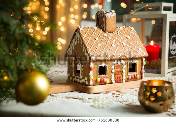 Little gingerbread house\
with glaze standing on table with tablecloth and decorations,\
candles and lanterns. Living room with lights and Christmas tree.\
Holiday mood