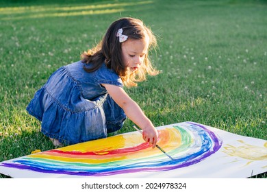 Little Girl 2-4 Years Old Paints Rainbow And Sun On Large Sheet Of Paper, Sitting On Green Lawn