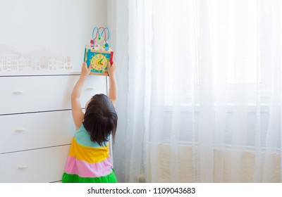 A Little Girl 3 Years Stretches To Get An Educational Toy From A High Shelf. Bright Dress On The White Background Of The Room. Free Space. Out Of Reach Of Children, Child Safety