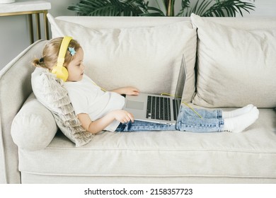 Little Girl 4 Years Old In Yellow Headphones In Front Of A Laptop. The Child Looks At The Computer Screen. The Baby Watches Cartoons While Mom Is Busy. Remote Learning For Preschool Children.