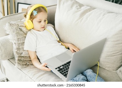 Little Girl 4 Years Old In Yellow Headphones In Front Of A Laptop. The Child Looks At The Computer Screen. The Baby Watches Cartoons While Mom Is Busy. Remote Learning For Preschool Children.