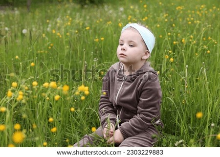 A little girl, 4 years old, sits in the big grass. A child on an uncut lawn. A child is playing on the street. Tall green grass in summer. Childhood in nature. Interested girl