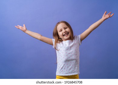 Little Girl Of 5 Years Old With Blonde Hair In A T-shirt With Colored Polka Dots Spreads Her Arms To The Side And Smiles Happily On A Purple Background. Close-up