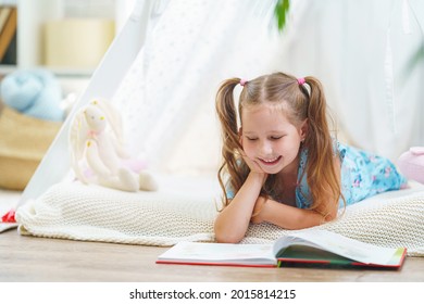 little girl 5 years old in blue dress, reading book with pleasure and looking at pictures with interest, lying in children's toy wigwam, child is at home. child's fantasy, happy childhood. Copy space