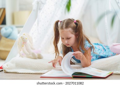little girl 5 years old in blue dress, reading book with pleasure and looking at pictures with interest, lying in children's toy wigwam, child is at home. child's fantasy, happy childhood. Copy space