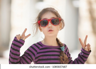A little girl of 6 years in sunglasses is happy and gesticulates with her hands and fingers - Shutterstock ID 1085837687