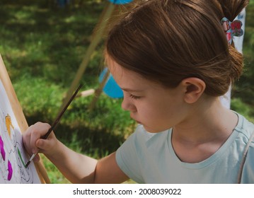 little girl 8-10 years old is painting on an easel diligently outdoors - Shutterstock ID 2008039022