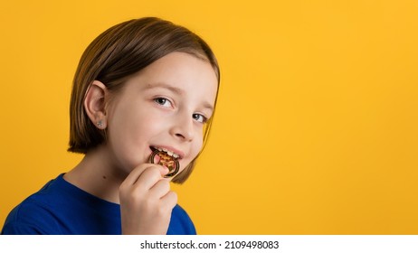 Little Girl 8-10 Years Old In Basic Blue T-shirt Biting Golden Physical Bitcoin On Yellow Background. Children Financial Literacy. Cryptocurrency And Blockchain Concept.