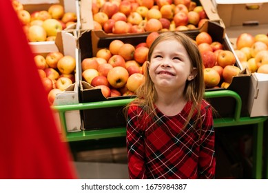 A Little Girl Of About 5 Threw A Tantrum In A Supermarket In Front Of Her Parents. The Child Screams And Cries, Begging Sweets From Mom And Dad On The Background Of Fruits And Apples